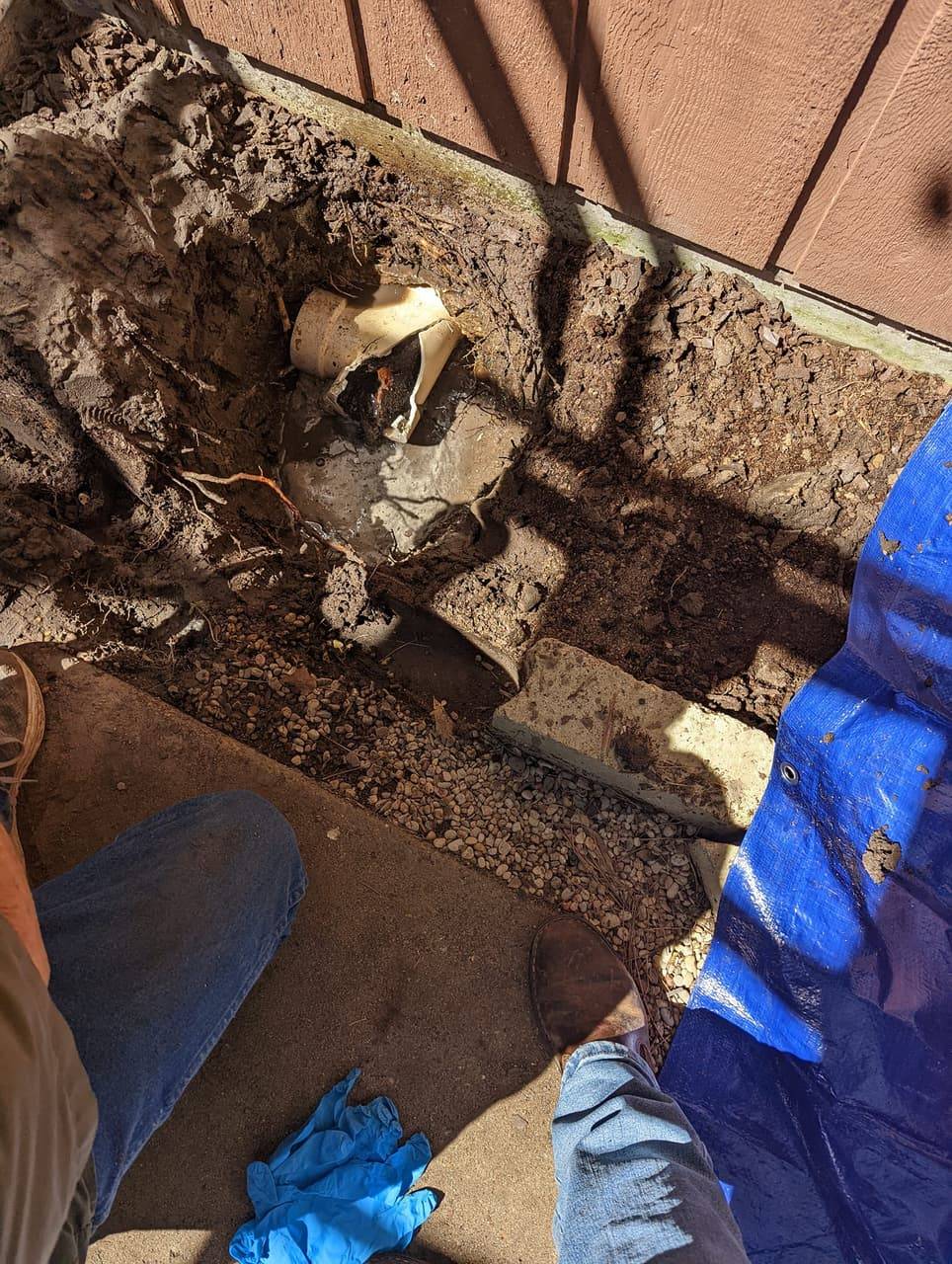 Excavated trench beside a house foundation exposing a broken drain pipe fitting